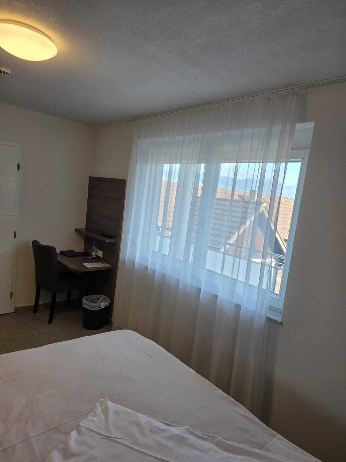 A hotel room with a bed, a desk, a chair, and a window with sheer curtains.