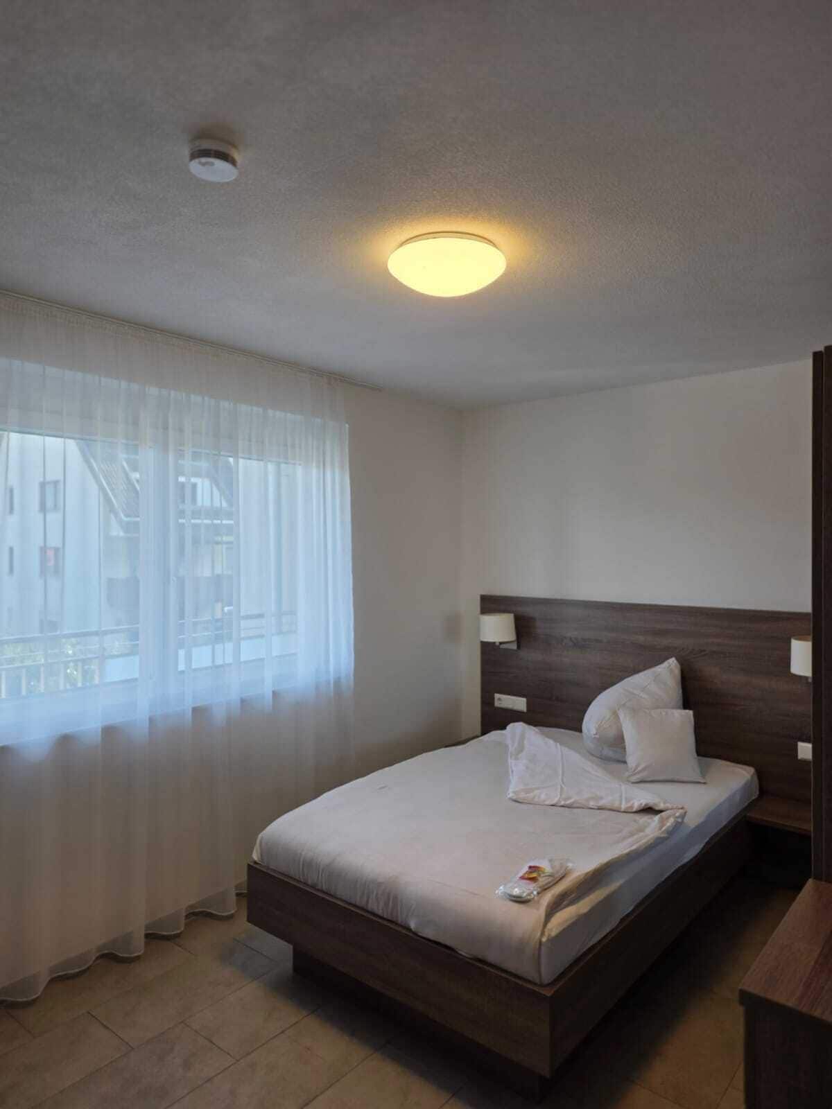 A hotel room with a bed, a bedside lamp, a window with sheer curtains, and a ceiling light.