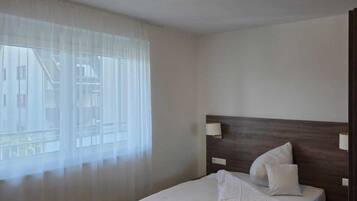 A hotel room with a bed, a bedside lamp, a window with sheer curtains, and a ceiling light.