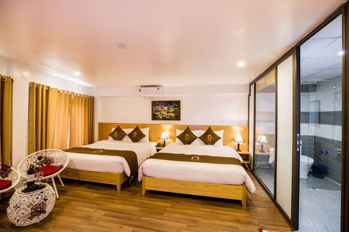 A hotel room with two beds, a sitting area with a chair and table, a painting on the wall, and a glass partition.