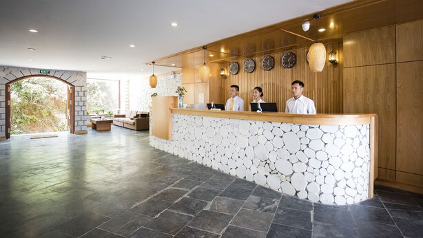 A reception area with a stone wall, wooden paneling, and a reception desk with three staff members. - BEAUTIFUL SAPA HOTEL (Sa Pa)