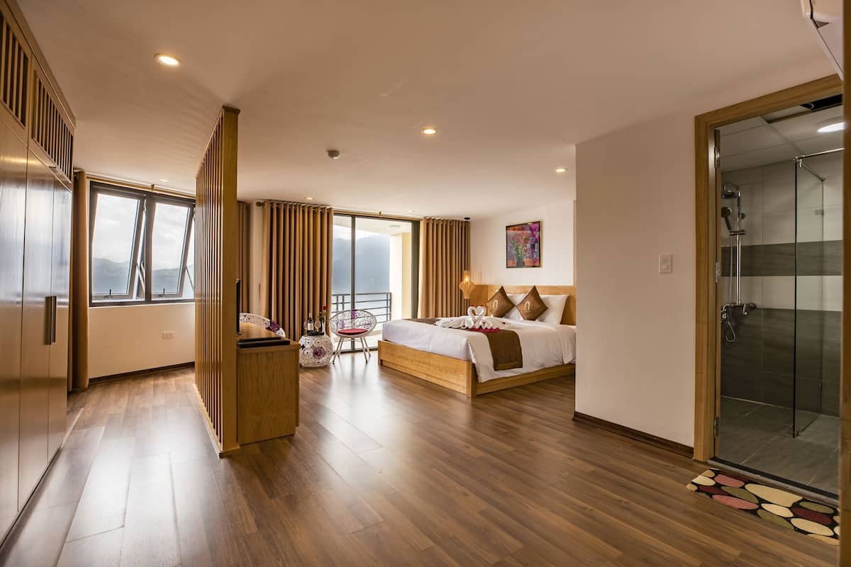 A modern hotel room with a large bed, a seating area with a chair and table, a glass shower enclosure, and a balcony with a view.