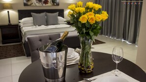 A hotel room with a bed, a dining table with a bottle of champagne, a vase with yellow roses, and a wine glass.