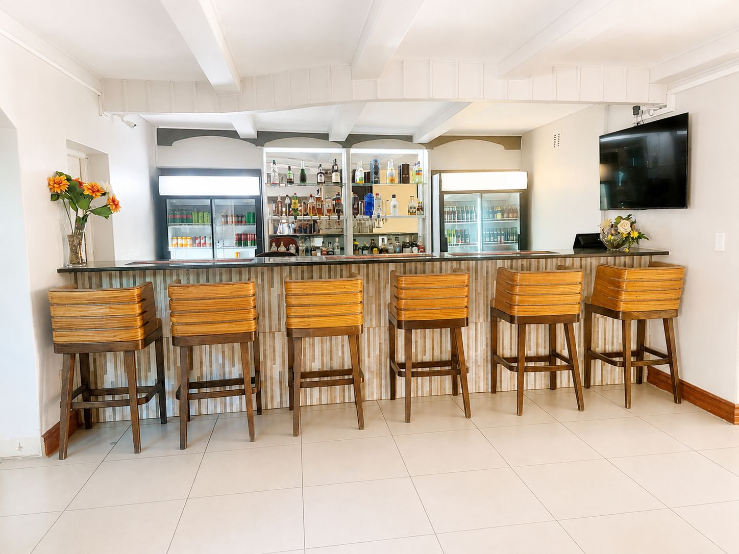 Eine Bar mit Holzbarhocker, einem mit Getränken gefüllten Kühlschrank und einem an der Wand befestigten Fernseher.