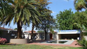 A pool lounge with a clear pool, palm trees, and a building with a sign. - Airport Garden Boutique Hotel (Boksburg)