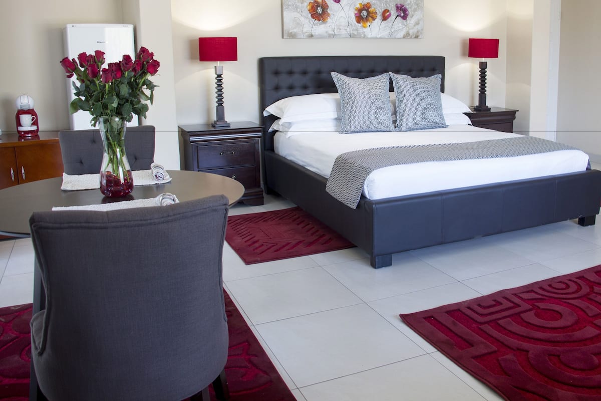 A modern hotel room with a large bed, two bedside tables, a nightstand, a round table with a chair, a vase with flowers, and a red rug.