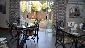 A dining area with a glass table and chairs, a view of the garden through glass doors, and a framed artwork on the wall.