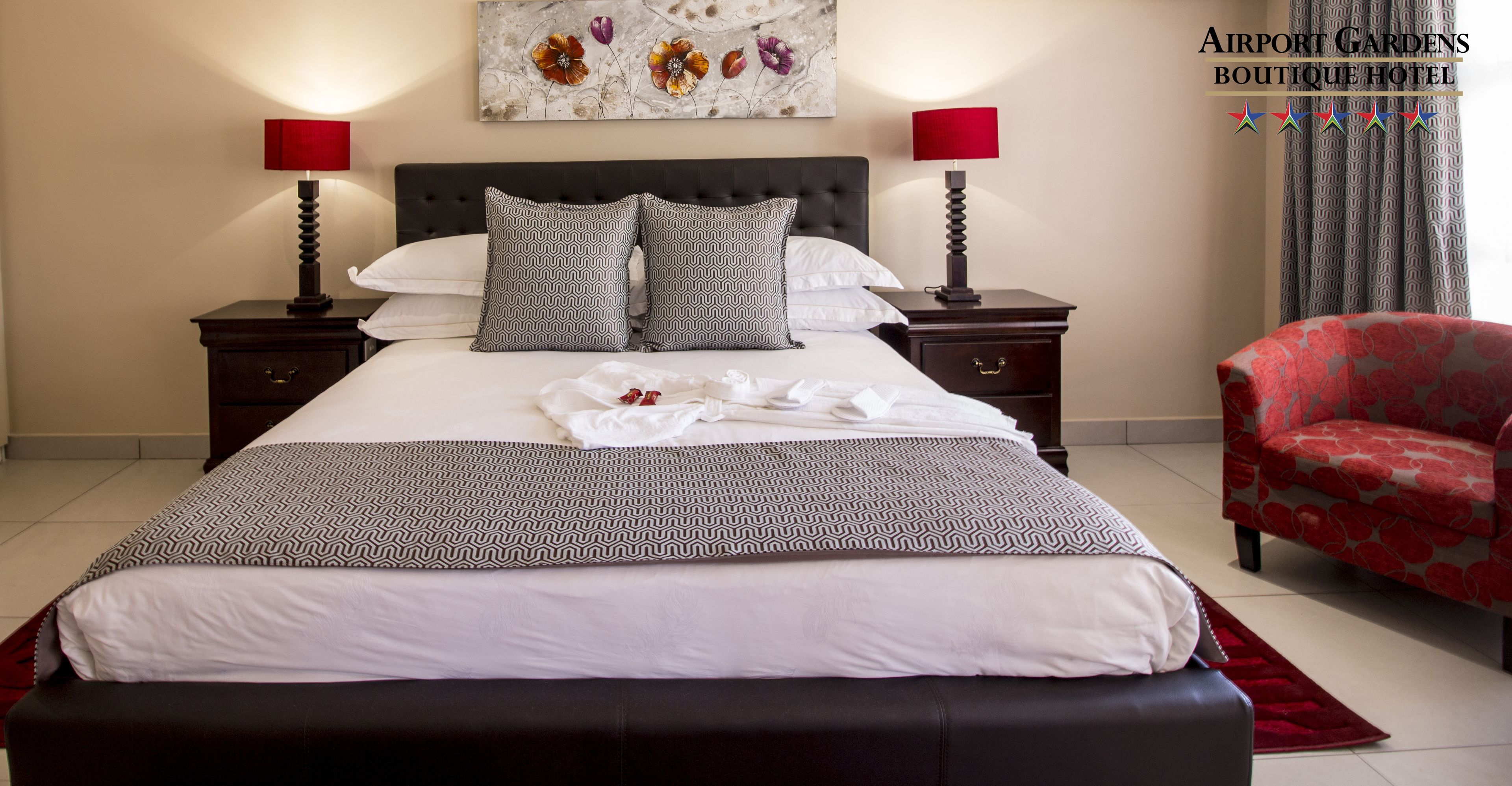 Cama bem arrumada com almofadas estampadas, mesa de cabeceira preta e vermelha, uma cadeira vermelha e uma obra de arte floral na parede.