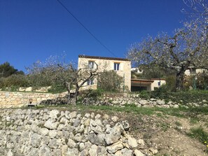 Uma casa de pedra com telhado de telhas, rodeada por um muro de pedra e vegetação.