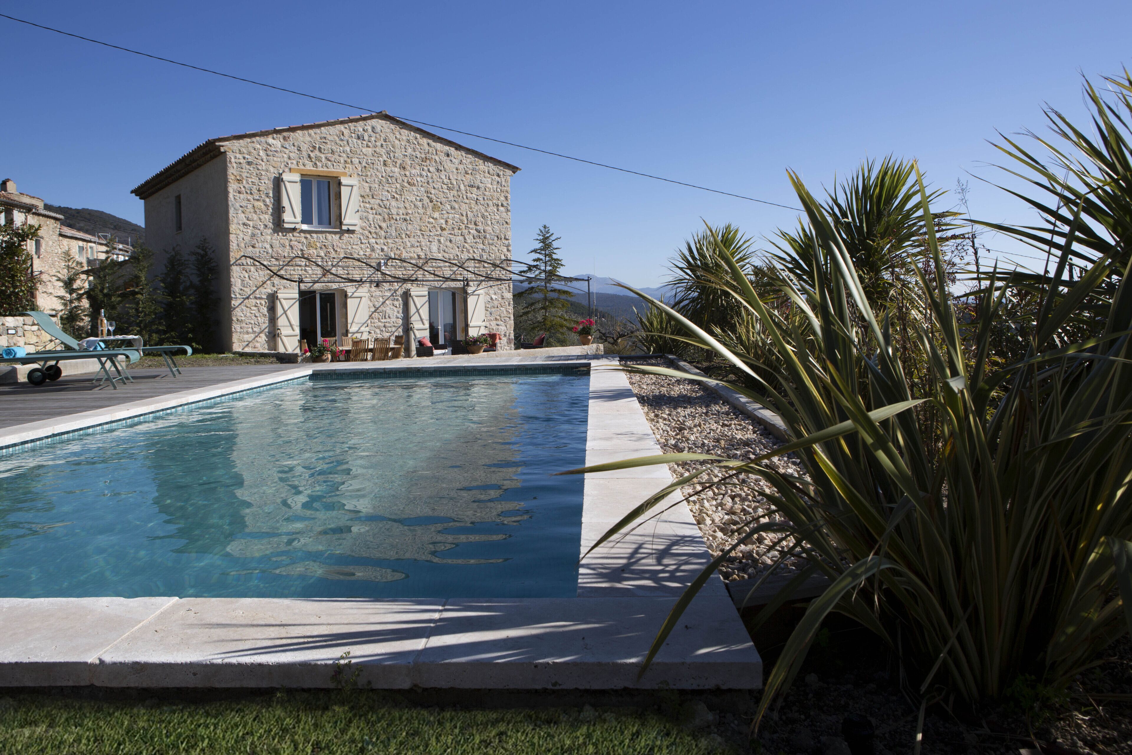 Una casa de piedra con piscina, rodeada de plantas y un cielo despejado.