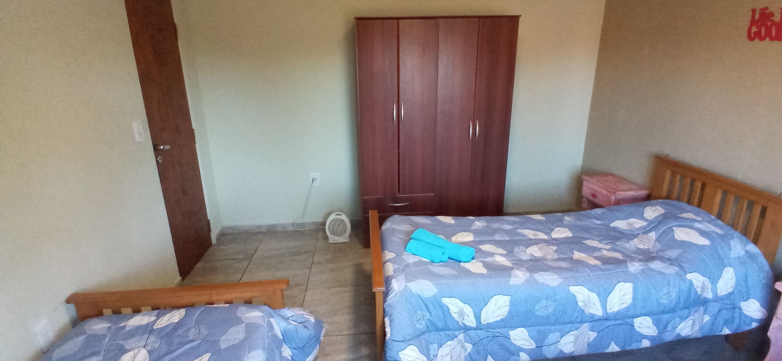 A bedroom with two beds, a wardrobe, and a small fan on the floor.