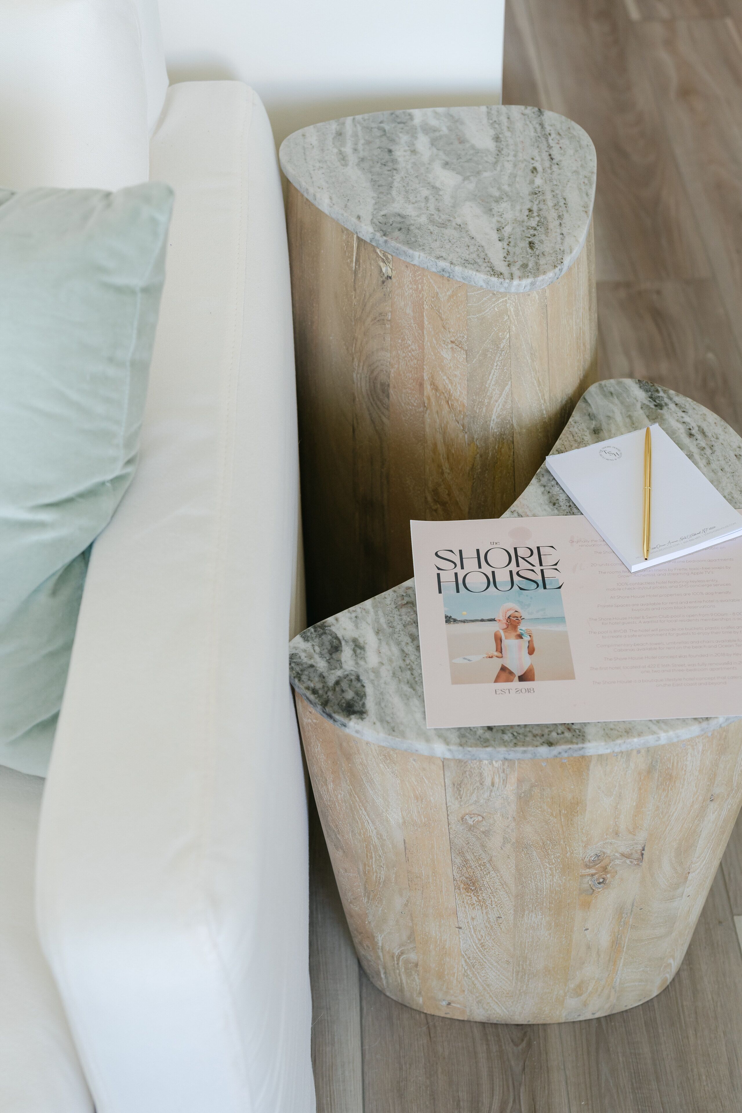 A white sofa with a light blue pillow, a wooden side table with a marble top, and a magazine titled "The Shore House" on the table.
