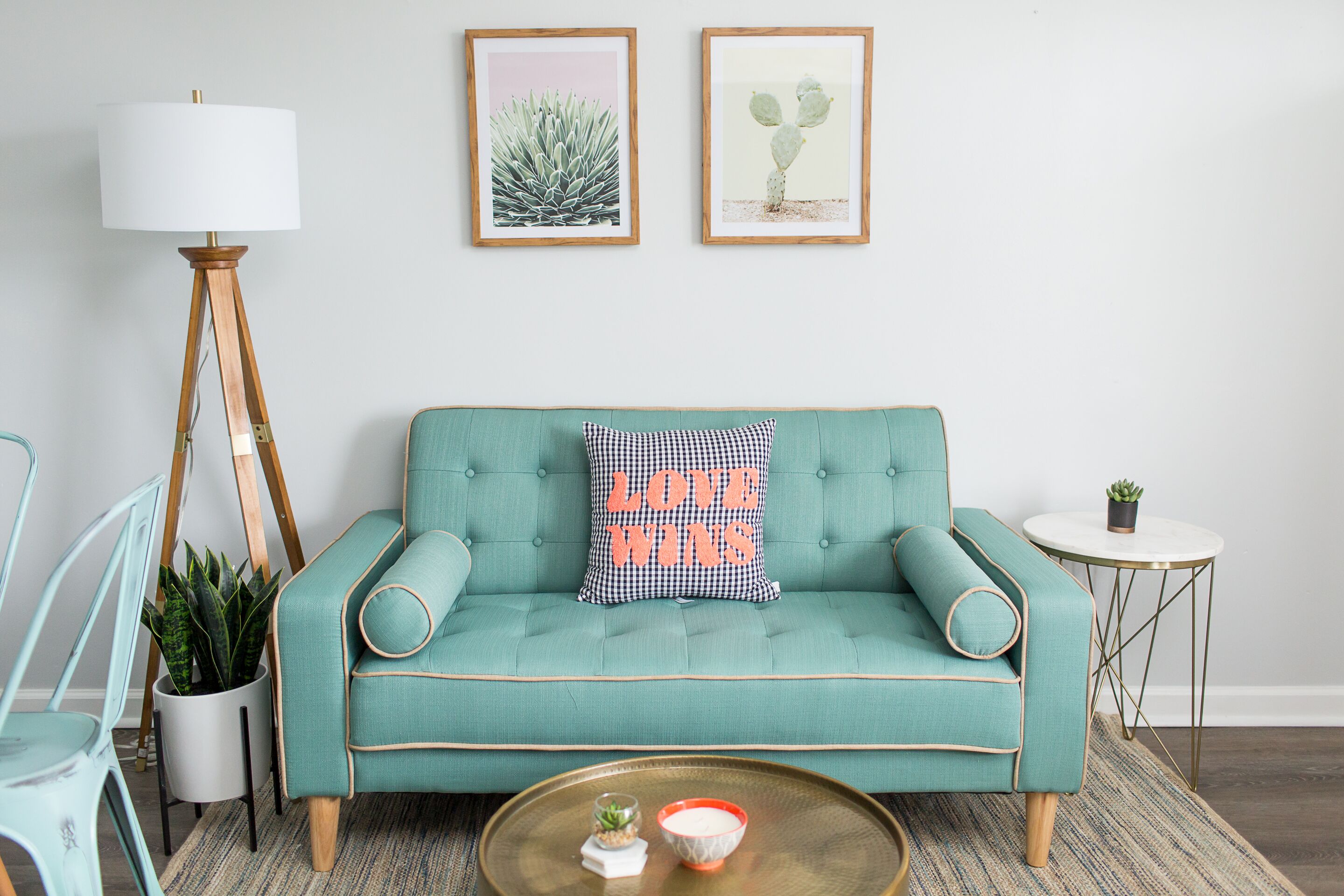 A teal sofa with a cushion that reads "LOVE WINS," a small round table with a plant, and two framed plant artworks on the wall.