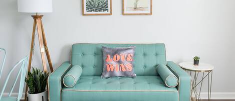 Un sofá color turquesa con un cojín que dice "LOVE WINS", una pequeña mesa redonda con una planta, y dos obras de arte con motivos vegetales en la pared.