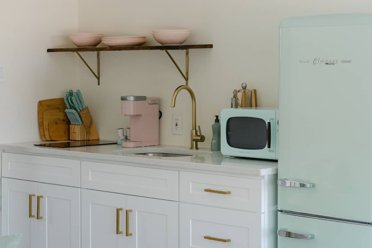 Una cocina con armarios blancos, una cafetera rosa, un microondas de estilo retro en color aguamarina y una tabla de cortar de madera.