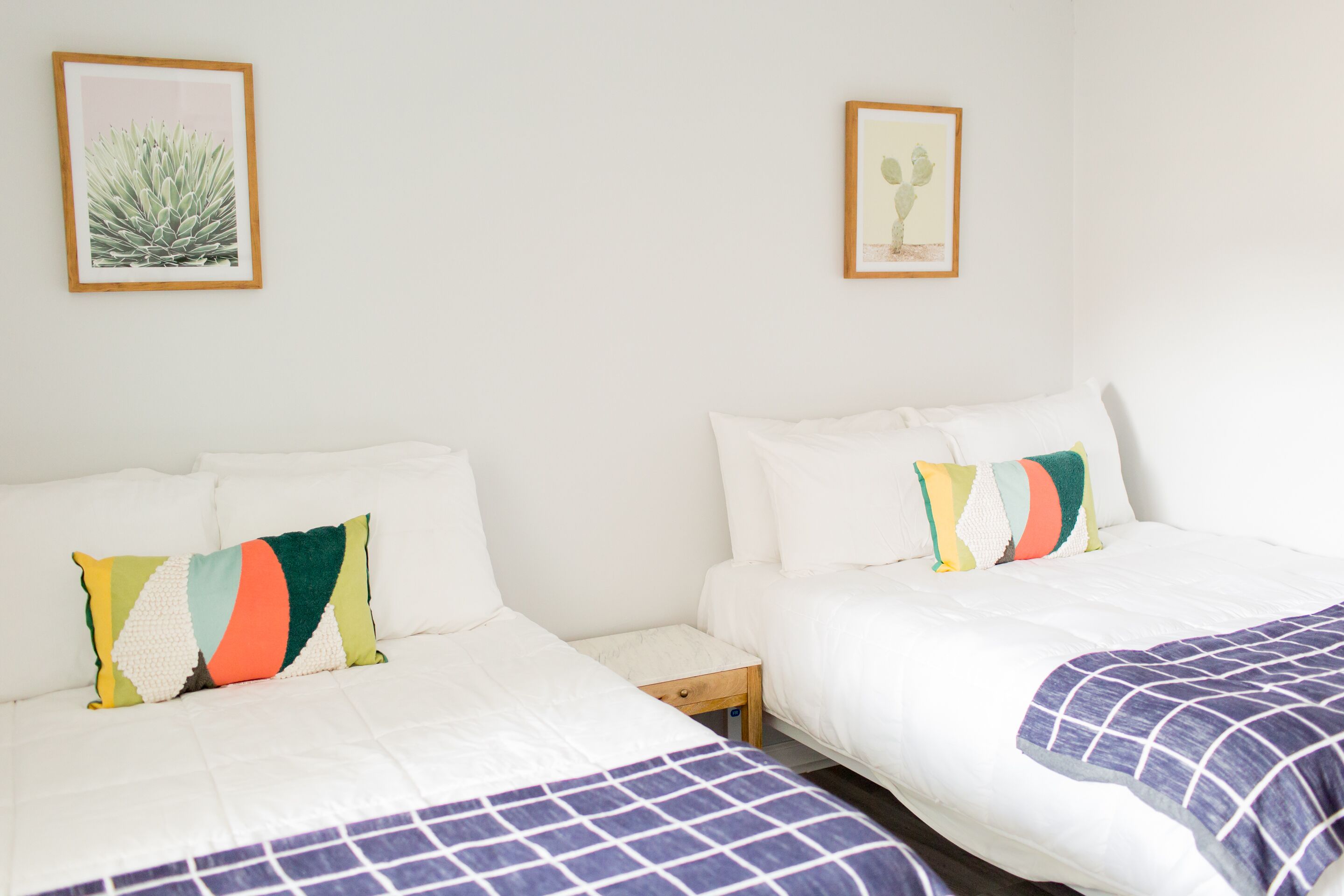 A hotel room with two beds, each with white bedding and colorful pillows. There are framed pictures on the wall above the beds.