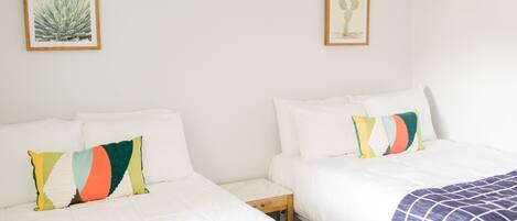 A hotel room with two beds, each with white bedding and colorful pillows. There are framed pictures on the wall above the beds.