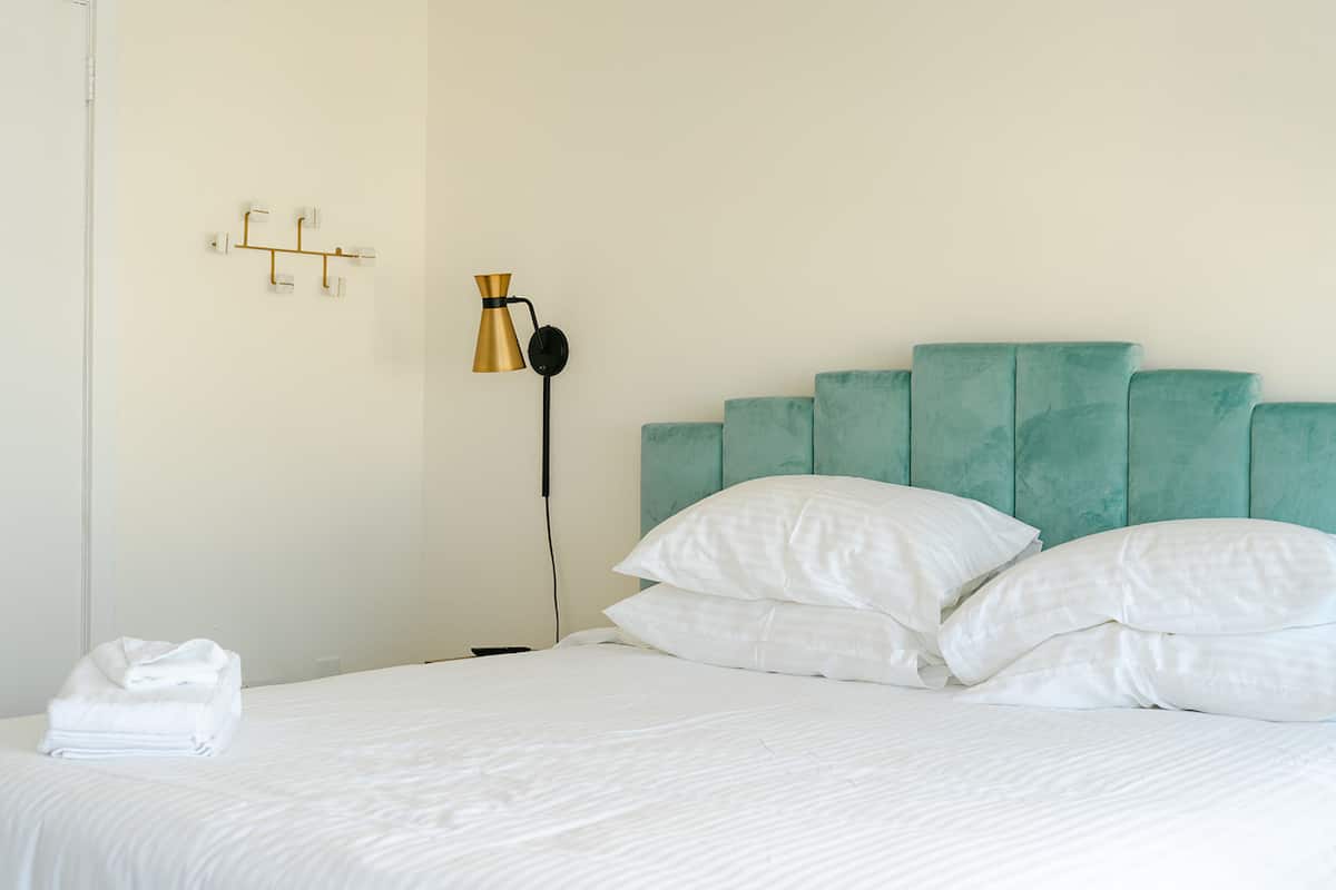 Una cama bien hecha con sábanas y almohadas blancas, un cabecero color turquesa y una lámpara de pared.