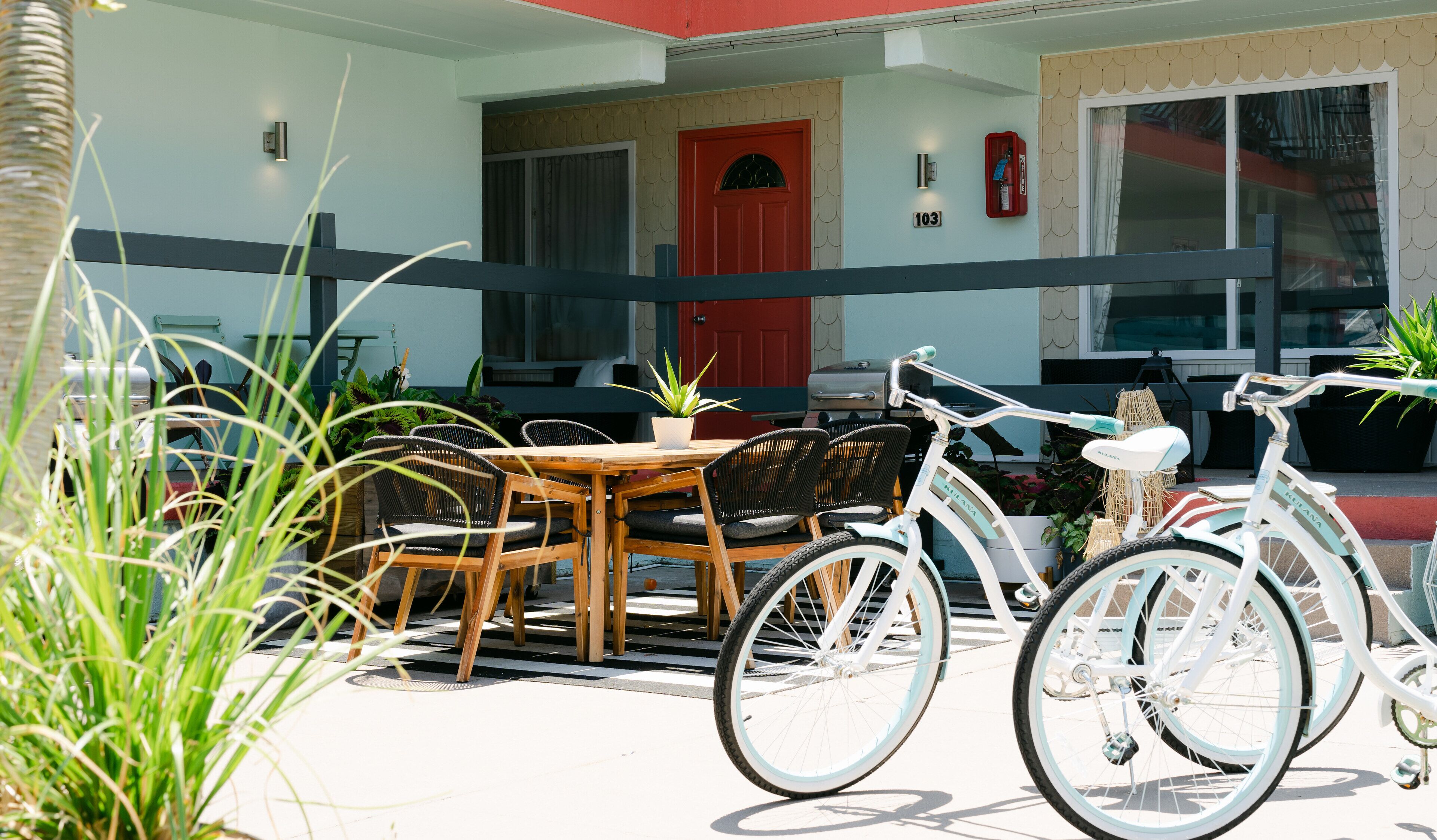 A patio area with a wooden table and chairs, a white bicycle, and a red door.