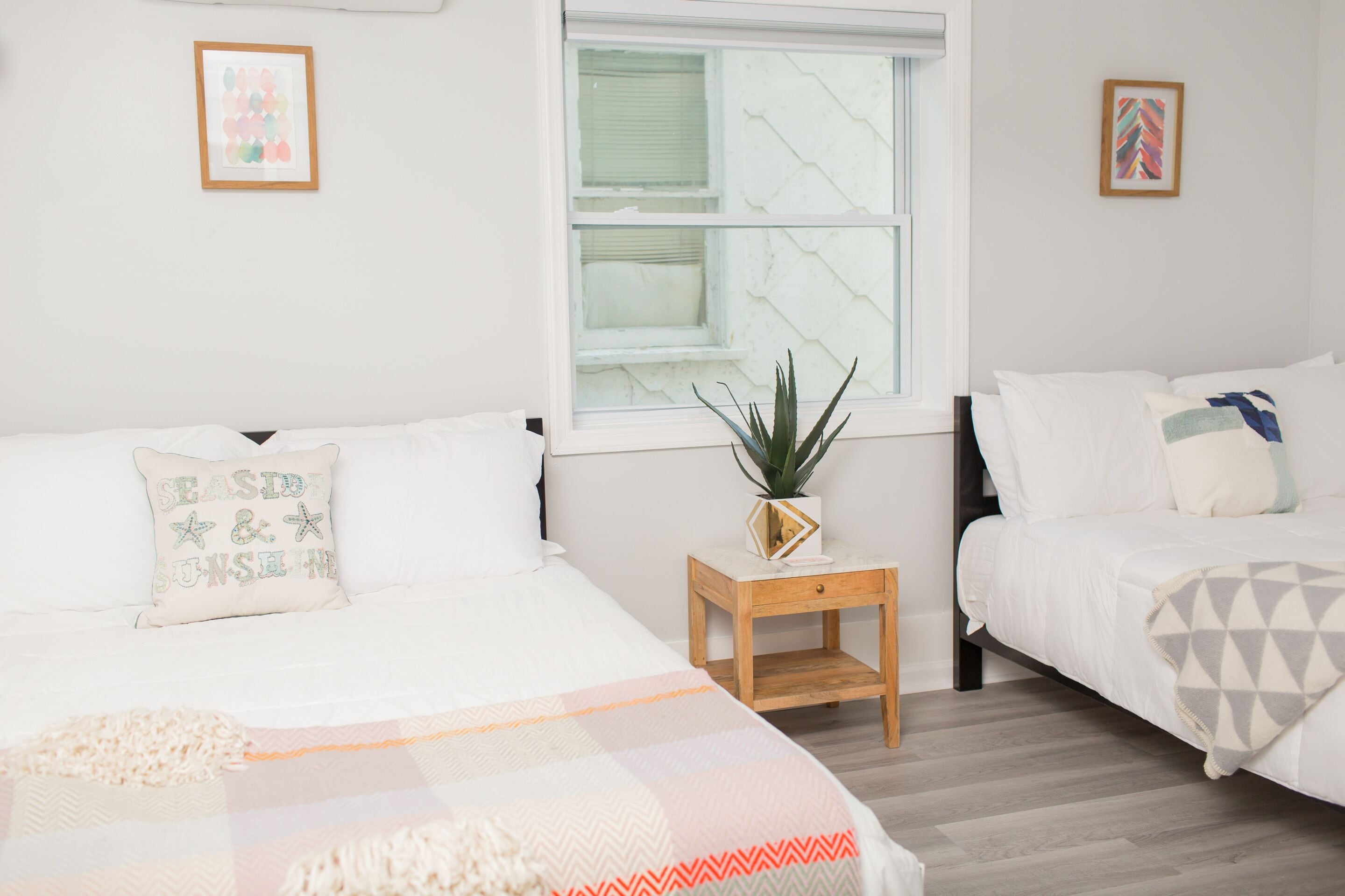 A bedroom with two beds, a window, and a small wooden nightstand with a plant.