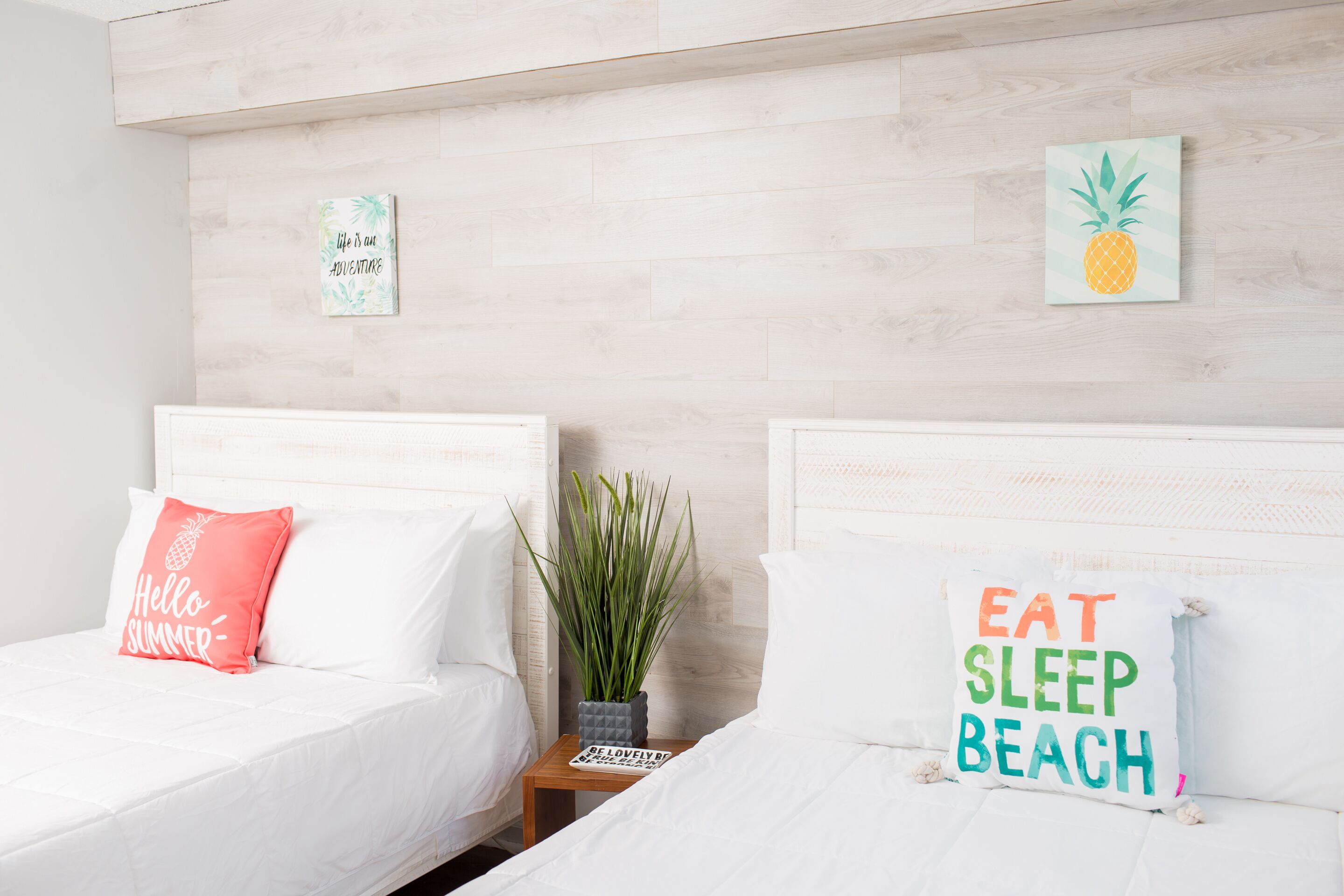 Habitación de hotel con dos camas, cada una con un almohadón decorado con motivos playeros.  Entre las camas, una pequeña mesa con una planta y un libro.