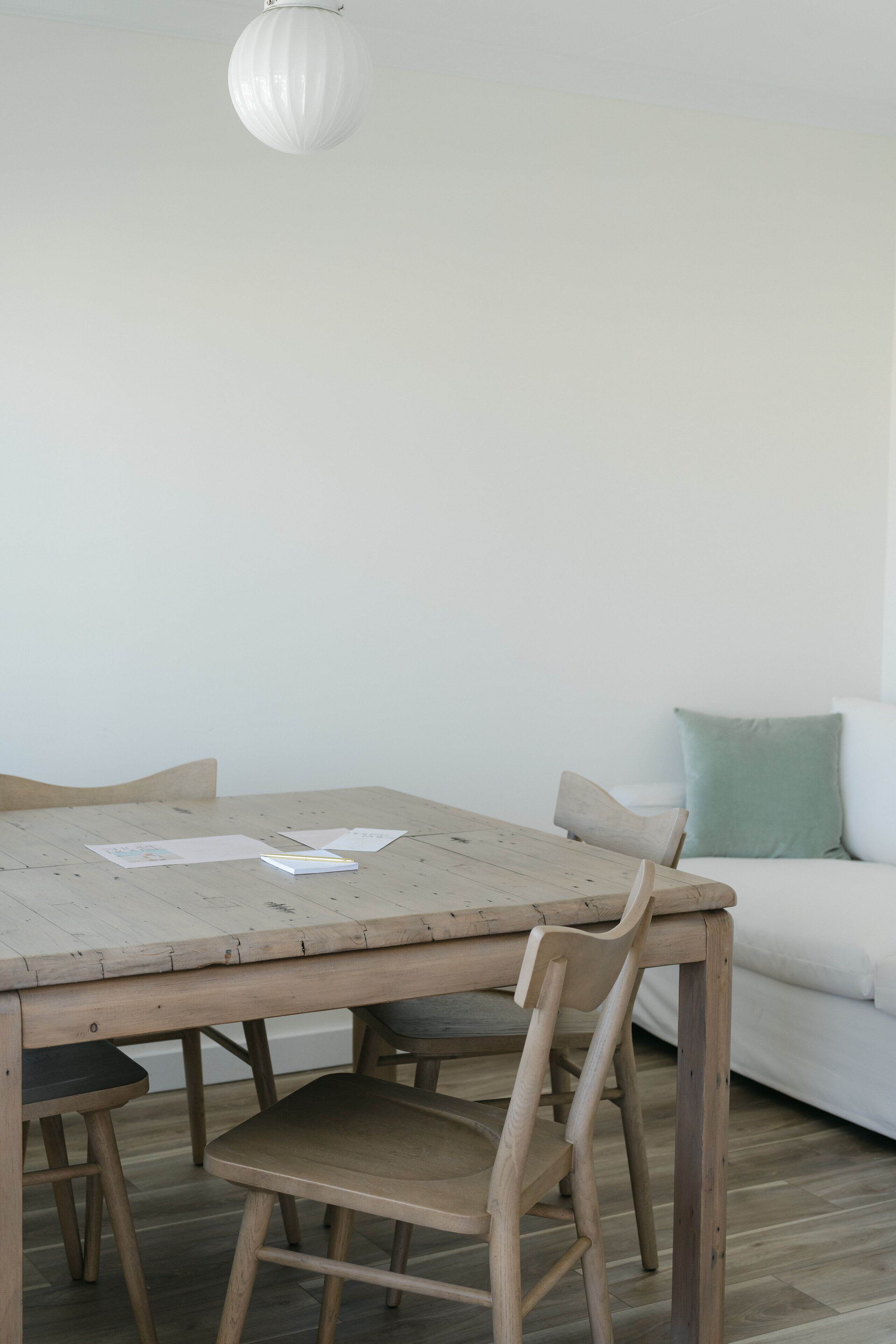 Un comedor con una mesa y sillas de madera, un sofá blanco con un cojín verde y una lámpara de techo blanca.