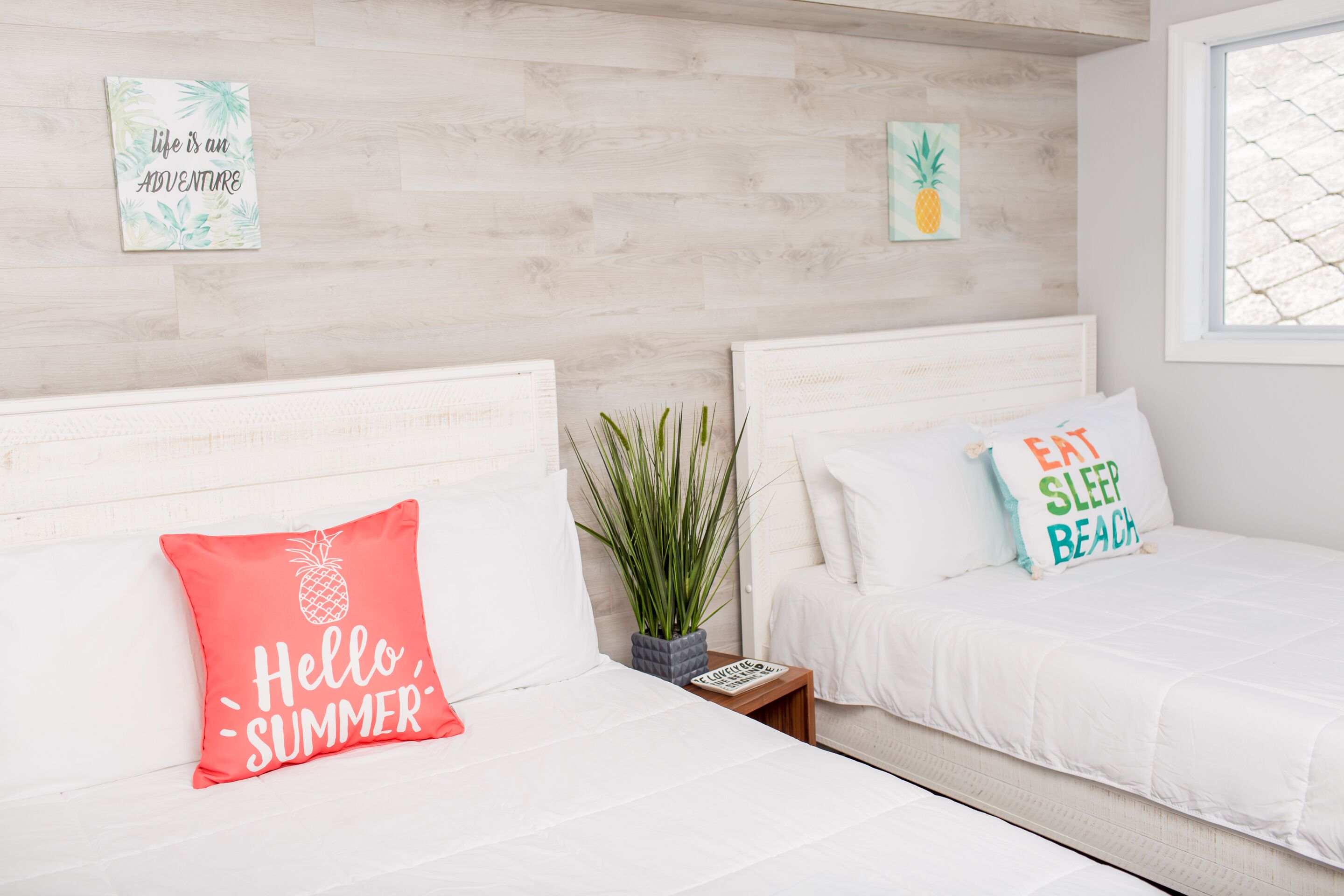 A bedroom with two beds, each with a different themed pillow, a small plant, and a wall with a pineapple artwork.