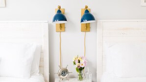 Una cama bien hecha con sábanas blancas, una mesita de noche con un reloj, un jarrón con flores y dos lámparas fijadas a la pared.