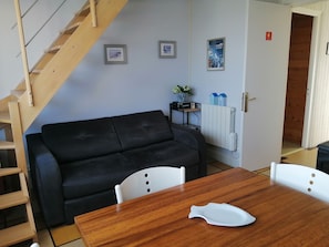 A living area with a black sofa, wooden dining table, white chairs, a wooden staircase, framed pictures on the wall, a radiator, and a door leading to another room.