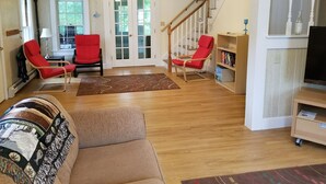 A living room with a wooden floor, two red chairs, a brown sofa, a ceiling fan, a bookshelf, and a staircase. - Charming Cape House in Megansett area of North Falmouth - walk to the beach! (North Falmouth)