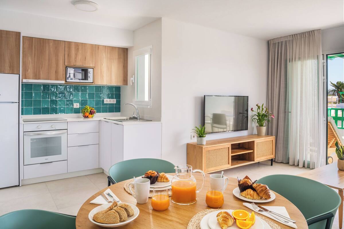 Eine moderne Küche mit weißen Schränken, einer Mikrowelle und einem Spülbecken. Es gibt einen Essbereich mit einem für das Frühstück gedeckten Tisch, inklusive Croissants, Orangensaft und einer Saftkanne.