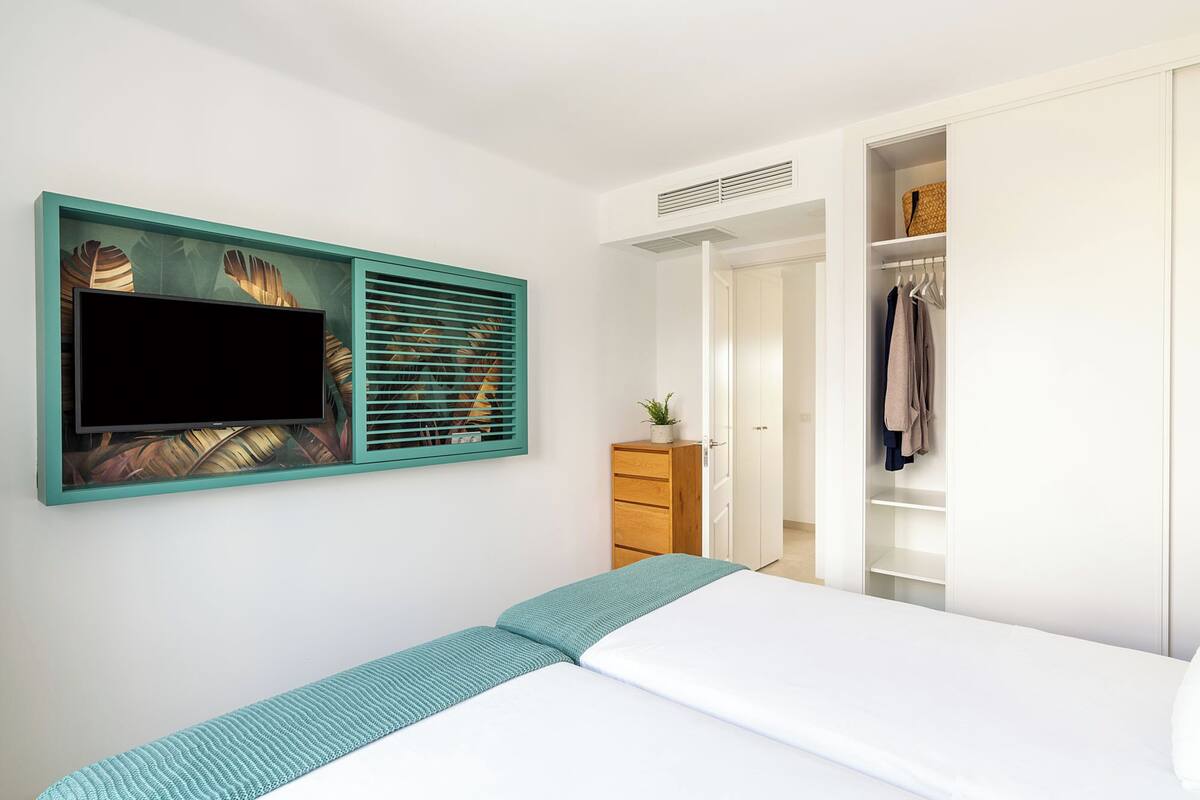 Ein Schlafzimmer mit einem Bett, einem an der Wand montierten Fernseher, einem Kleiderschrank und einer kleinen Pflanze auf der Kommode.
