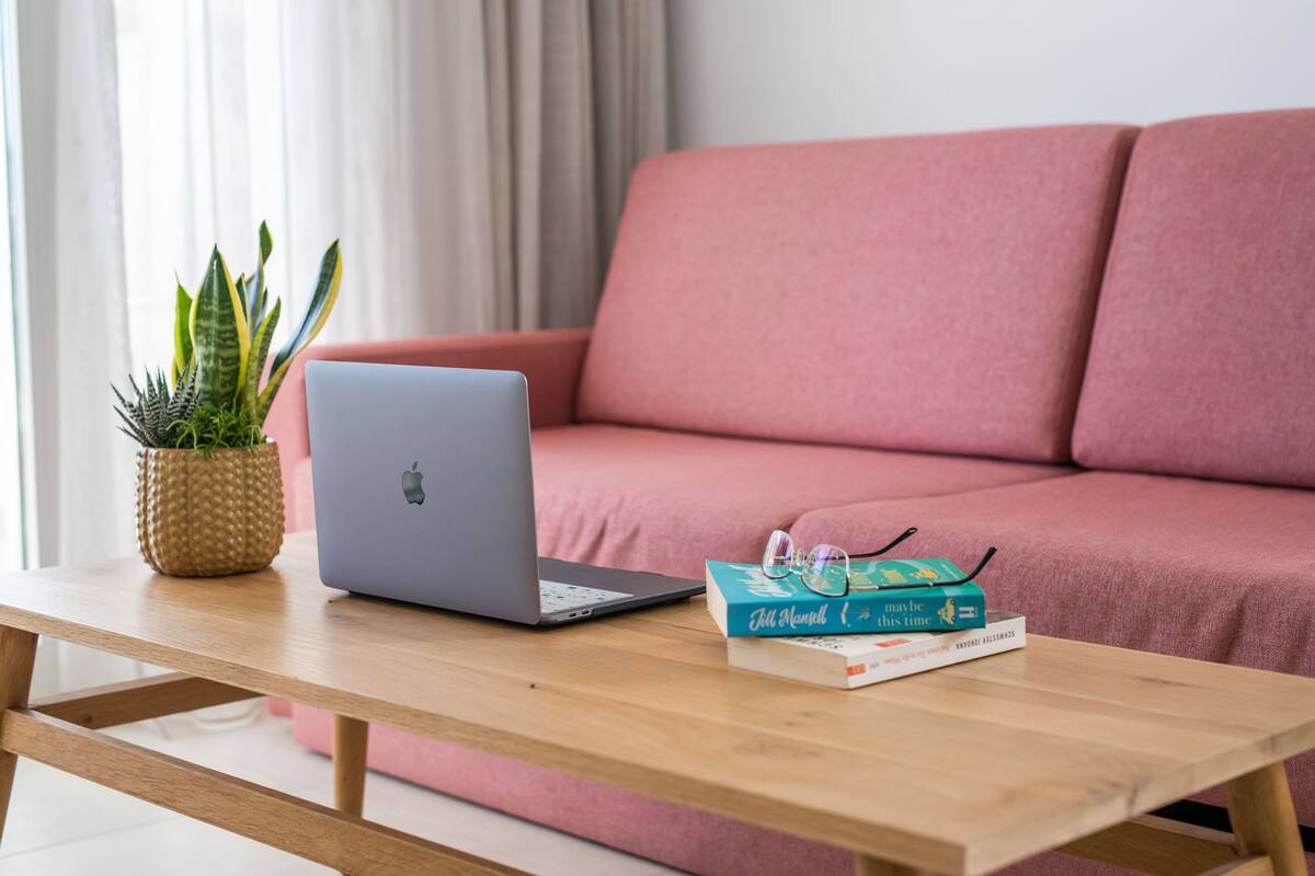 Ein Laptop auf einem Holztisch, umgeben von Büchern und Brillen, vor einer pinkfarbenen Couch.