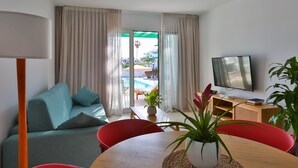 A modern living room with a sofa, red chairs, a wooden table, a TV, and a view of an outdoor pool area. - Nazaret Apartments (Teguise)