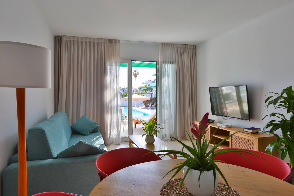 Ein modernes Wohnzimmer mit einer Couch, roten Sesseln, einem Holztisch, einem Fernseher und Blick auf einen Außenpool.