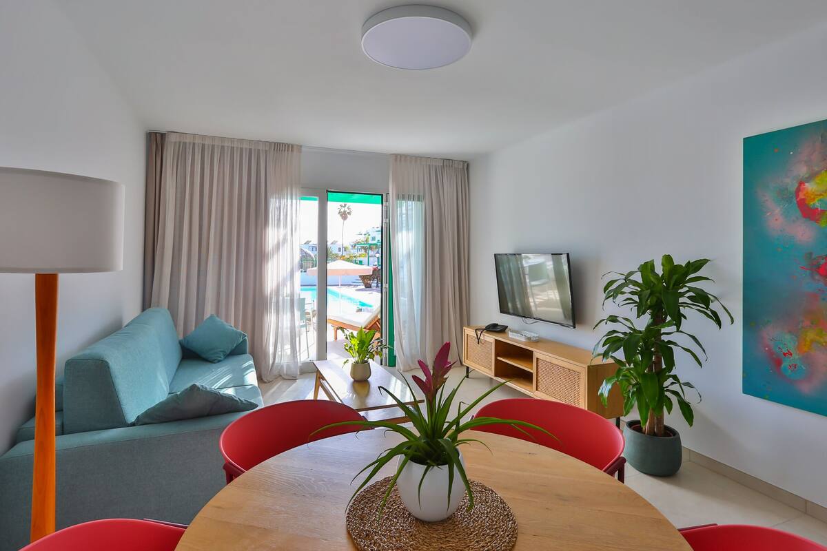 Ein modernes Wohnzimmer mit einer Couch, einem Esstisch, Stühlen, einem Fernseher und einem großen Fenster mit Vorhängen.