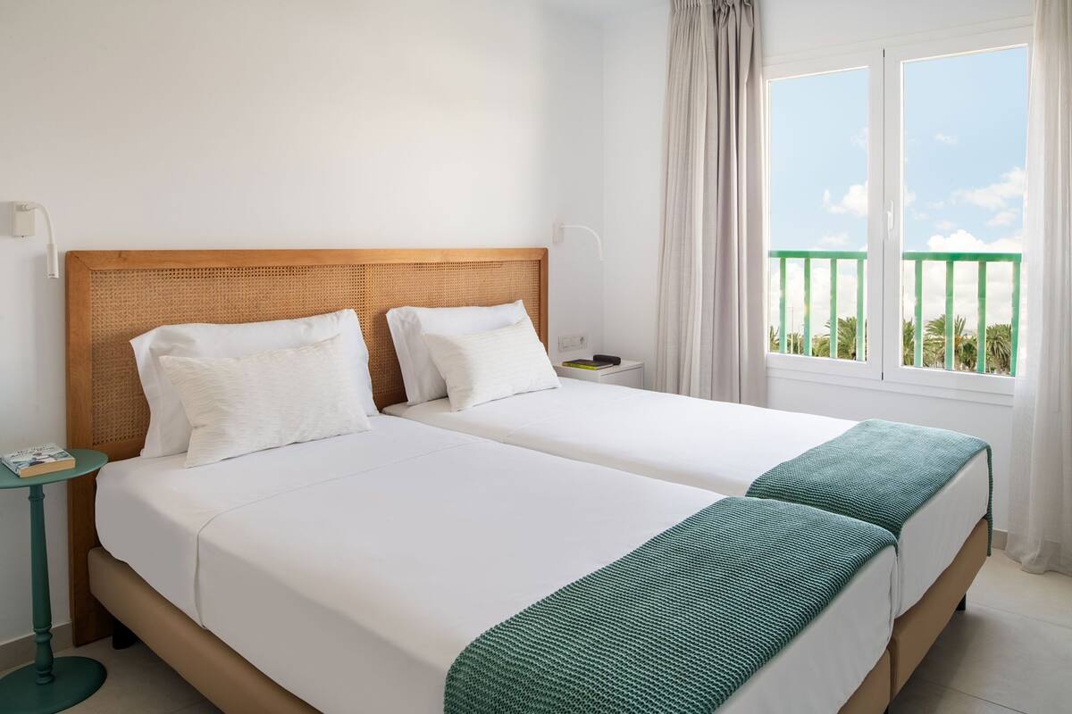 Ein Doppelbett mit weißer Bettwäsche und einer grünen Decke, ein hölzerner Kopfteil, ein Nachttisch mit einem Buch und ein Balkon mit grüner Brüstung.