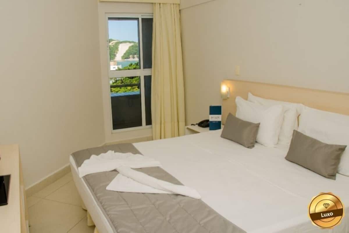 Ein Hotelzimmer mit Bett, Fernseher, einem Fenster mit Vorhängen und Blick auf Grünflächen.