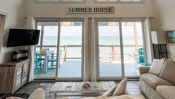 A spacious living room with a ceiling fan, a TV, and a sofa. The room has a large window and a door leading to a balcony with a view of the ocean. - Modern Beachfront Cottage with Endless Bay Views (Port Haywood)