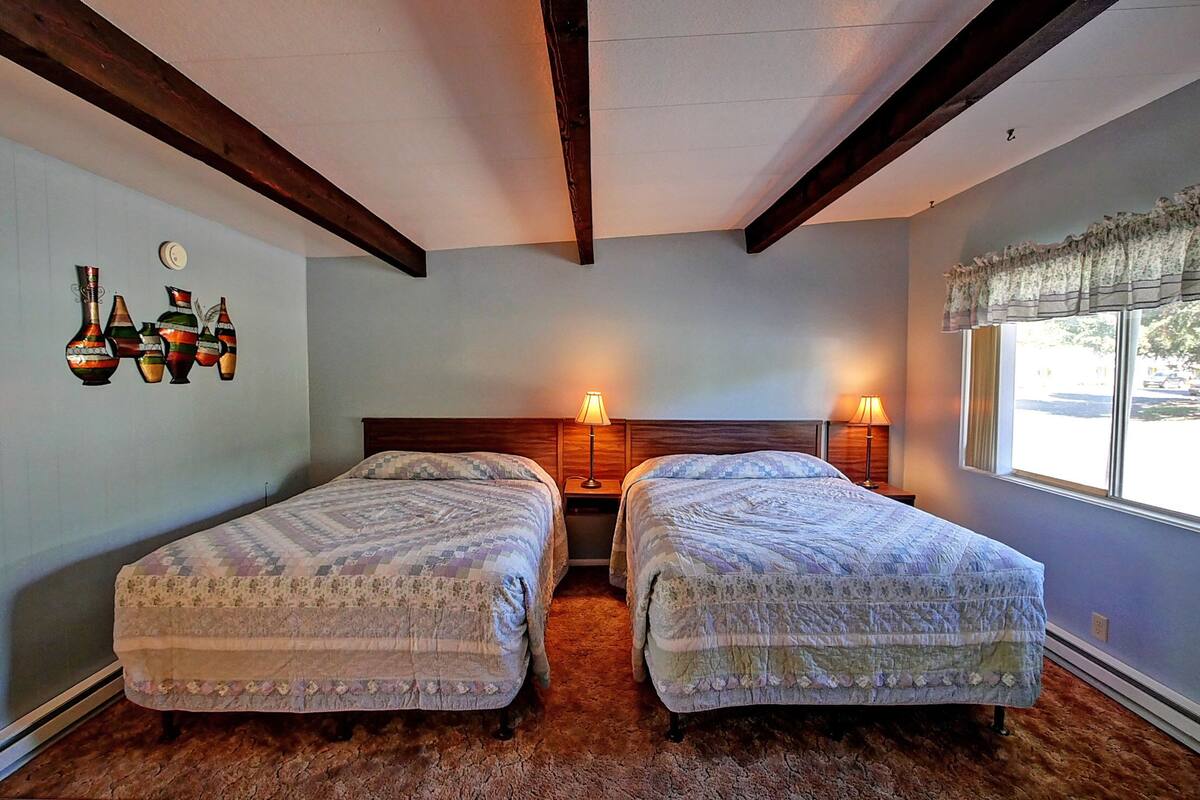 A bedroom with two beds, wooden beams on the ceiling, a window with a valance, and wall-mounted lamps.