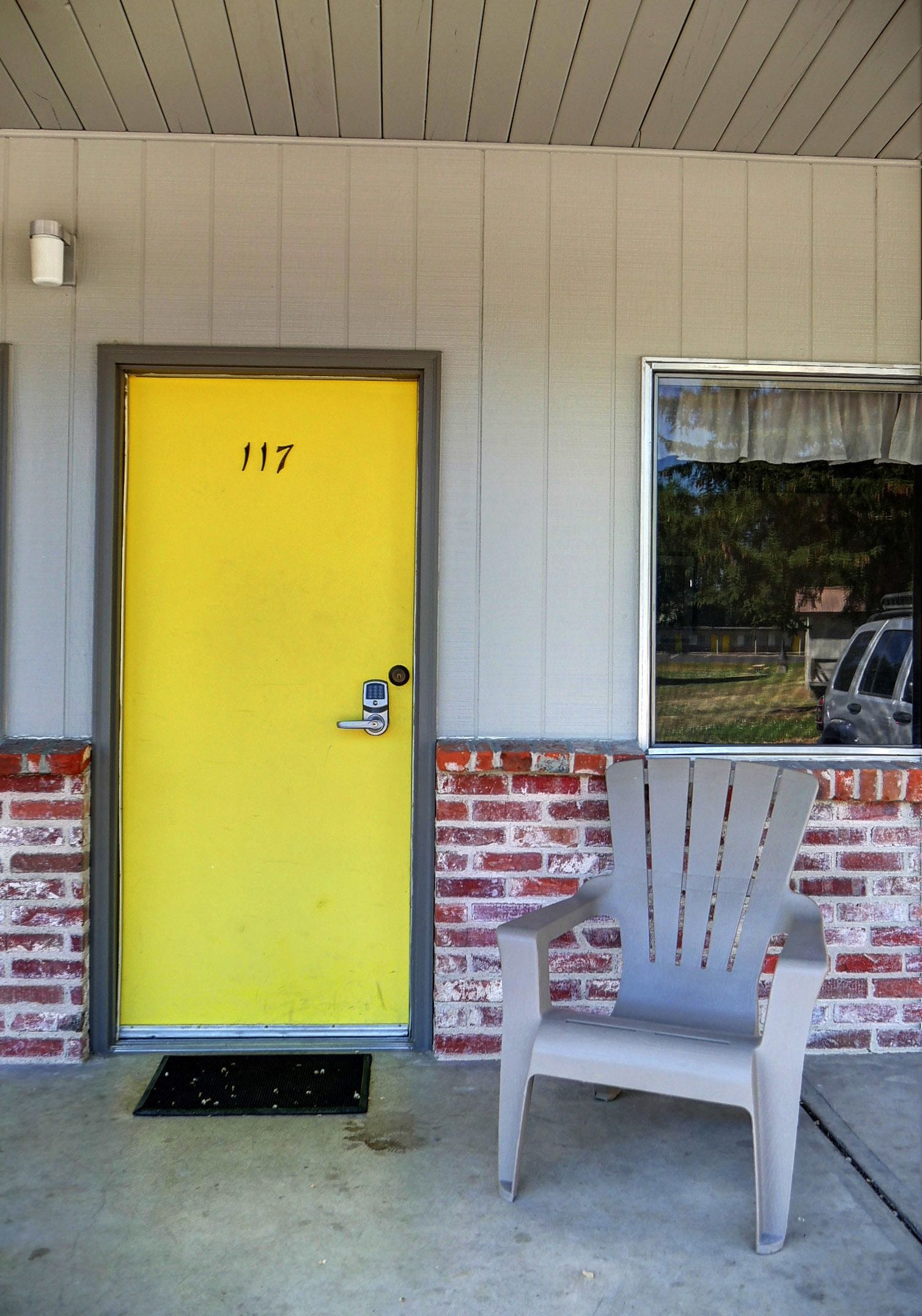 A yellow door with the number 117, a gray plastic chair, and a window reflecting a vehicle and trees.