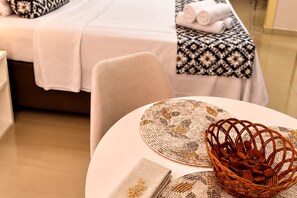 Une chambre d’hôtel avec un lit parfaitement fait, une table ronde avec un set de table et un panier d’amandes, et un chemin de table à motifs.
