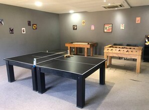 Una mesa de ping pong, una mesa de futbolín y una pequeña mesa de madera en una habitación con paredes grises.