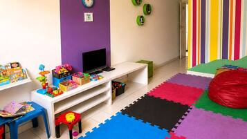 Uma sala de brincar infantil com um móvel branco para entretenimento, brinquedos coloridos e um grande puff.