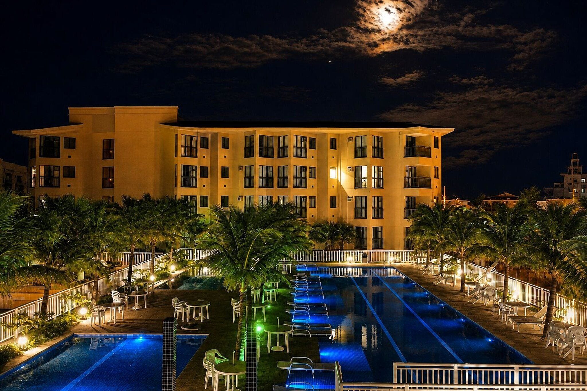 Hotel com piscina, palmeiras e céu noturno.