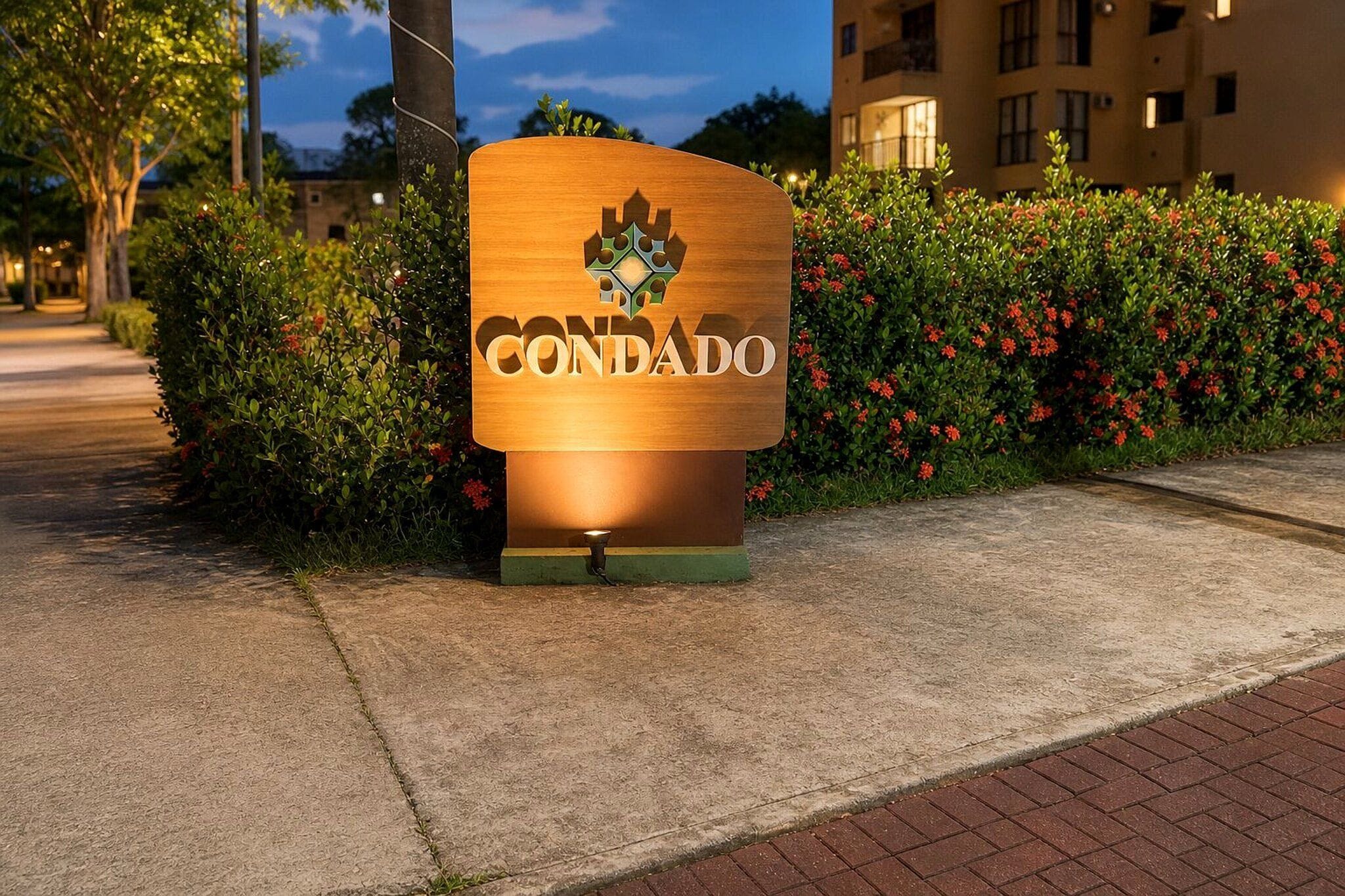 Uma placa de madeira com a palavra 'CONDADO' e um logotipo, colocada em uma calçada ao lado de um arbusto com flores vermelhas.