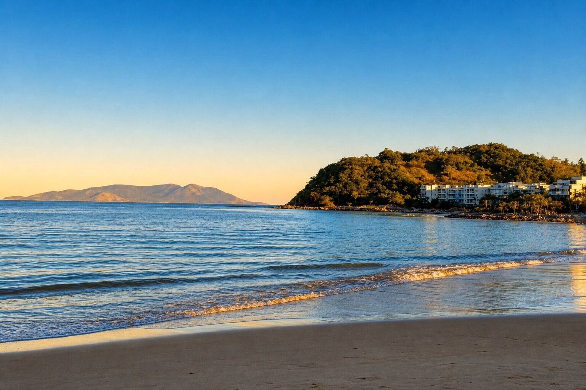 Uma praia com vista clara para o mar, areia dourada e uma colina com edifícios ao fundo.