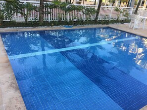 Una piscina azul cristalina con un borde empedrado, rodeada por una cerca blanca y follaje.