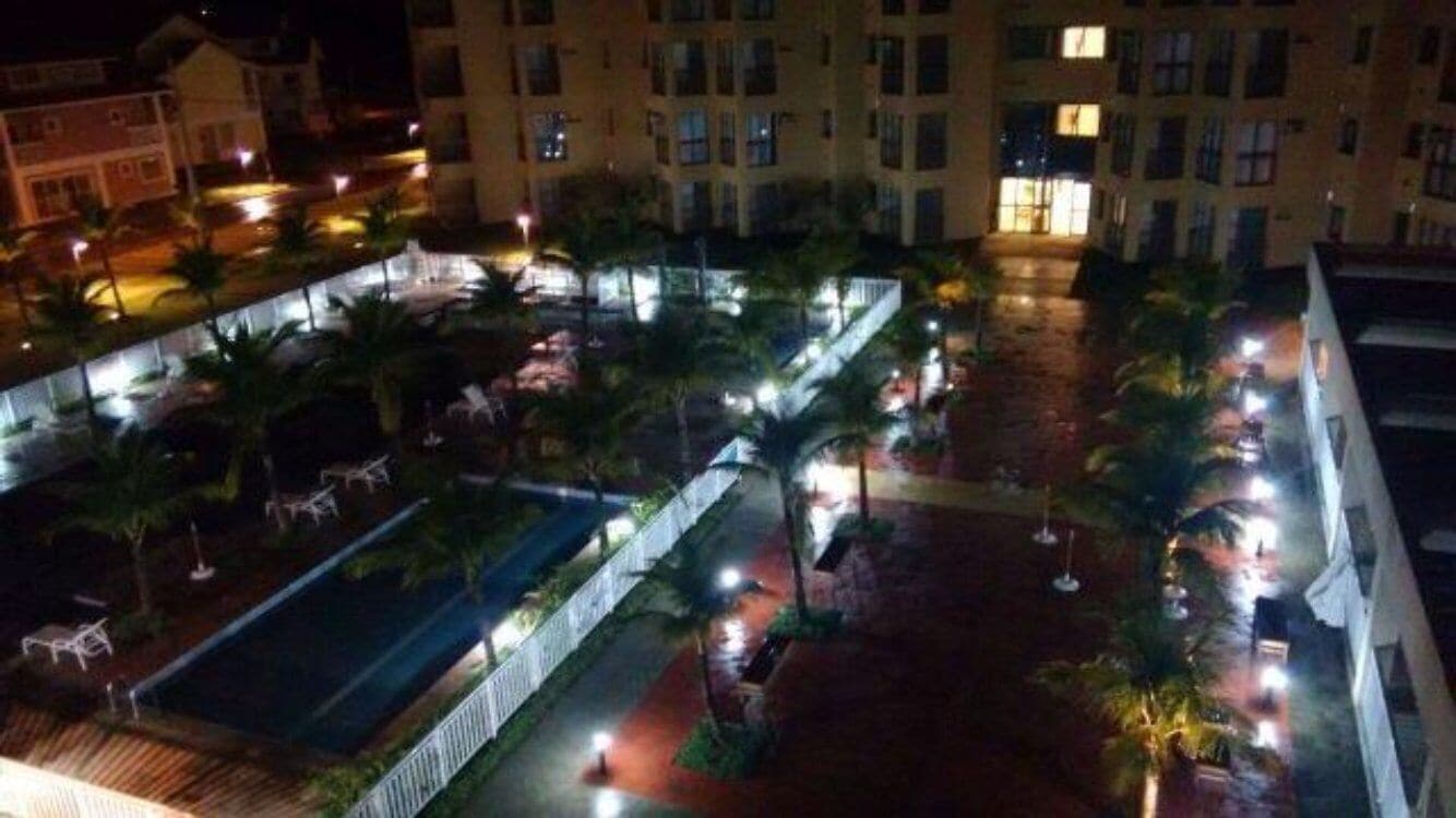 Vista noturna de um pátio de hotel, com palmeiras, piscina e um caminho bem iluminado.