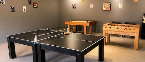 A room with a ping pong table, a foosball table, and various posters on the walls.